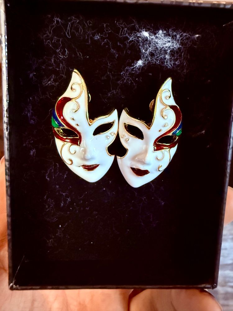 A black box containing two white Venetian mask like faces with fancy red gold and greens filagree around one of their hallow eyes. Their red lips are upturned in very slight smiles