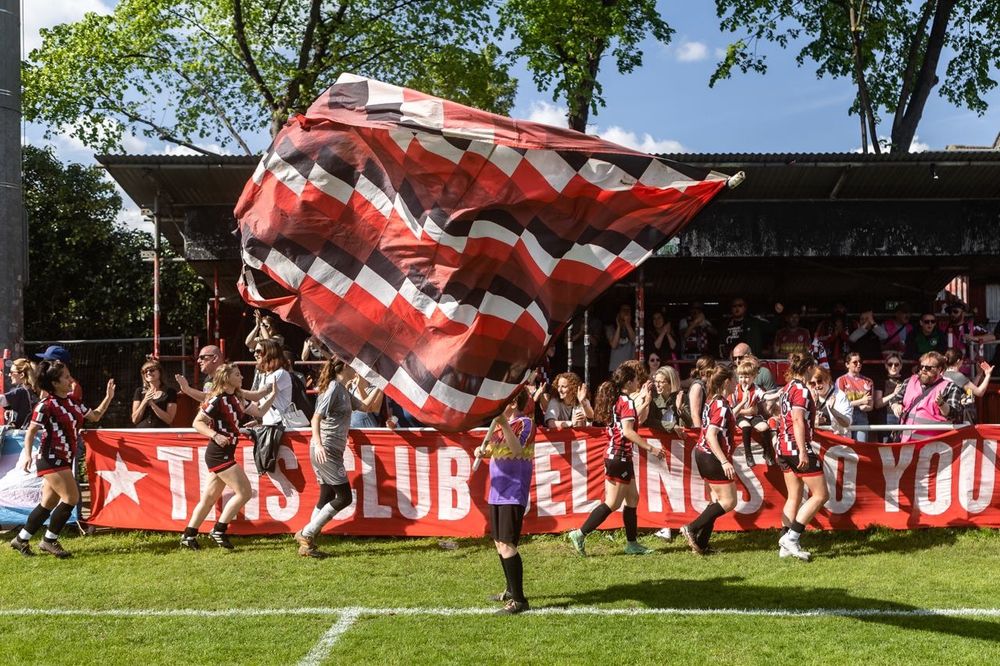 The Clapton CFC Women’s First Team celebrating with home fans under the ‘scaffold’ stand at the Old Spotted Dog Ground.