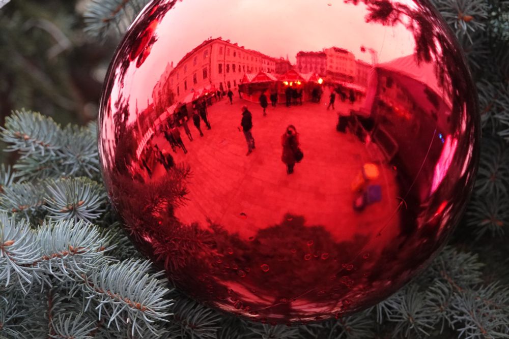 A red sphere in a green pine tree reflecting me taking a photo and a "rounded" landscape of an old European town square with some Christmas market stalls wet up and a handful of people walking around.