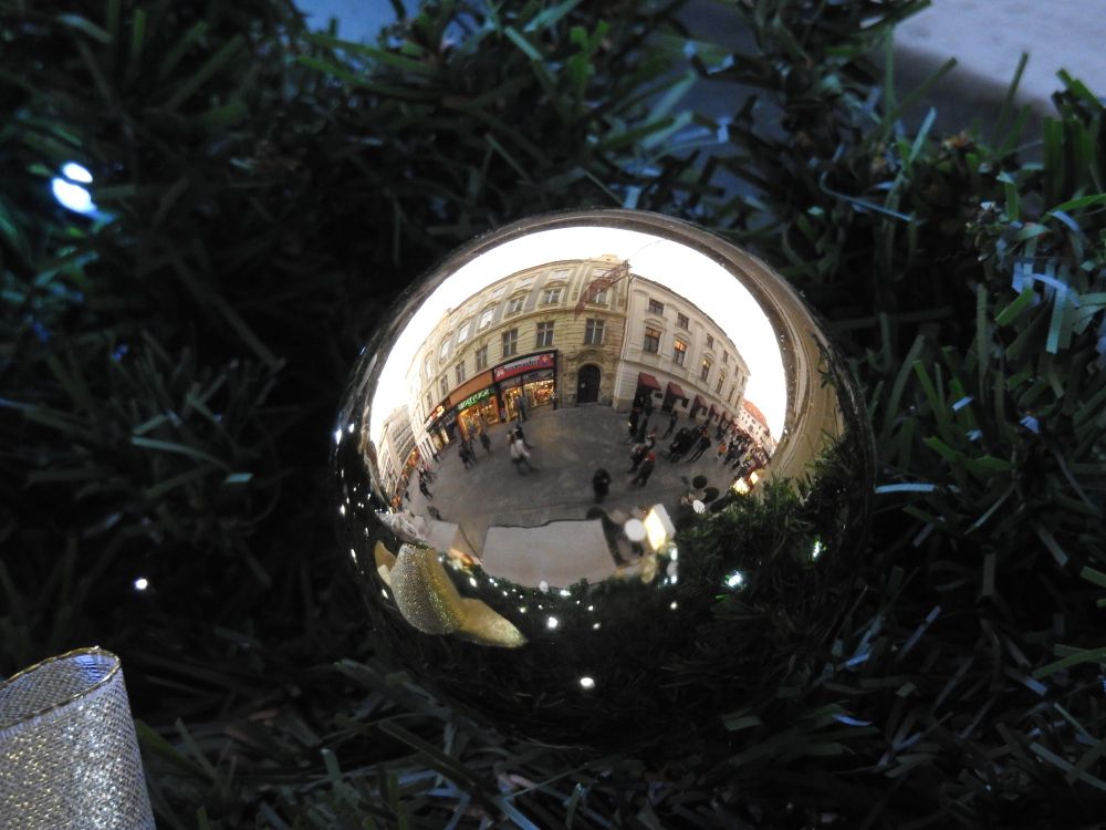 A white/beige Christmas ornament that is reflecting back two old buildings with storefronts and the pedestrian crowd. Because it is daylight, the reflection is quite sharp and although the street is rounded in the shape of the sphere, details like the shop windows and people walking by are quite visible.