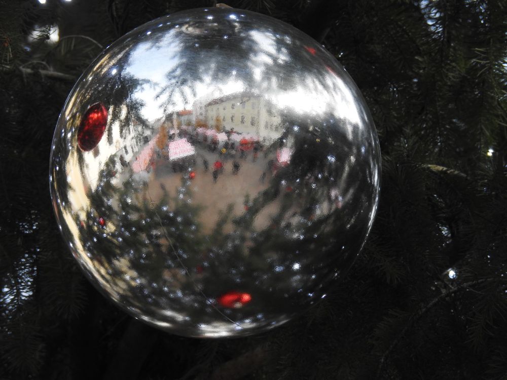 A white/mirror Christmas ball reflecting back some of the tree it is sitting in, as well as some of the other red ornaments. A dayscape of rows of red and white market stalls appear to be peeking through the leaves in the reflected scene.