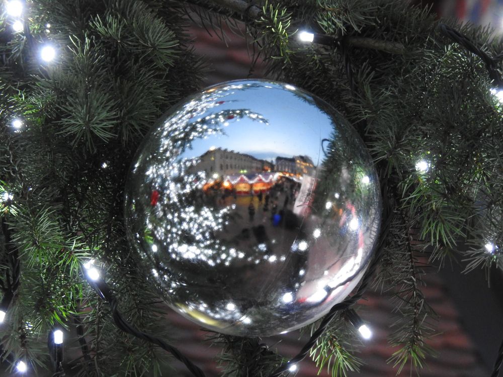 A dusky, evening scene reflected in a white Christmas ball. The tree has strings of tiny white lights which are reflected back in the ball, and beneath them a lit up old European town square with Christmas market stalls all in a row, joined by lights are visible.