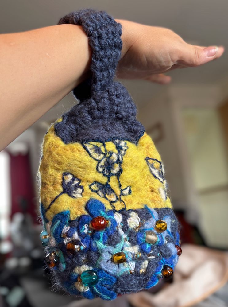 Small wrist bag hanging from a white arm. The bag has a crocheted top with a strap that goes round the wrist, the body of the bag is tubular with a yellow felted top embroidered with blue flowers that have shading in felt of blue and white. The bottom half is felted in the same blue as the crocheted top and covered felted and embroidered flowers and and tiny insects with beads as part of the design.