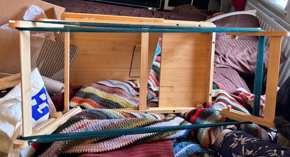Heap of blankets with various bags and bits of thread jumbled up in it. A folding table designed for sitting in bed is lying on its side with the tilting top used as a prop, around the legs is wound green warp thread.
