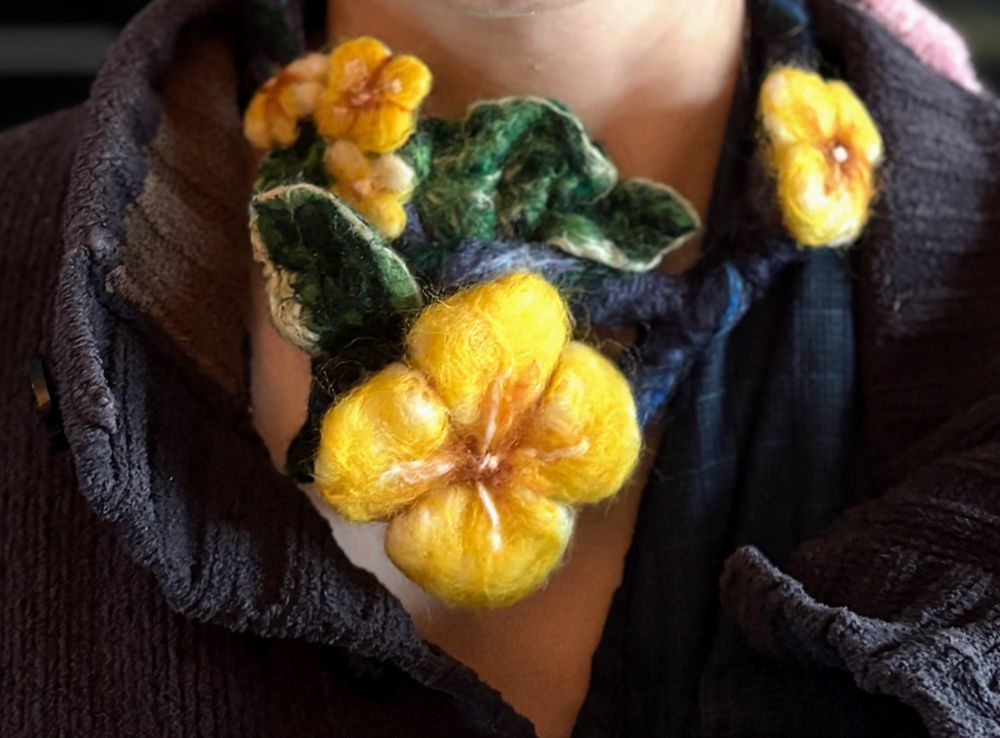  A circle of felted material in swirls of blue with five yellow flowers and two leaves, that forms a necklace. The necklace is pictured on a disembodied white neck.