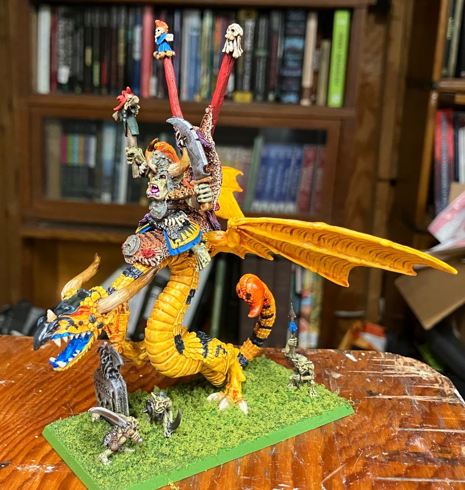 Left view of a bright yellow wyvern with black stripes and a red-tipped tail, being ridden by an orc wearing a huge horned helmet and carrying a sword and puppet. The model is on a bright green base covered with model railroad flock. 
