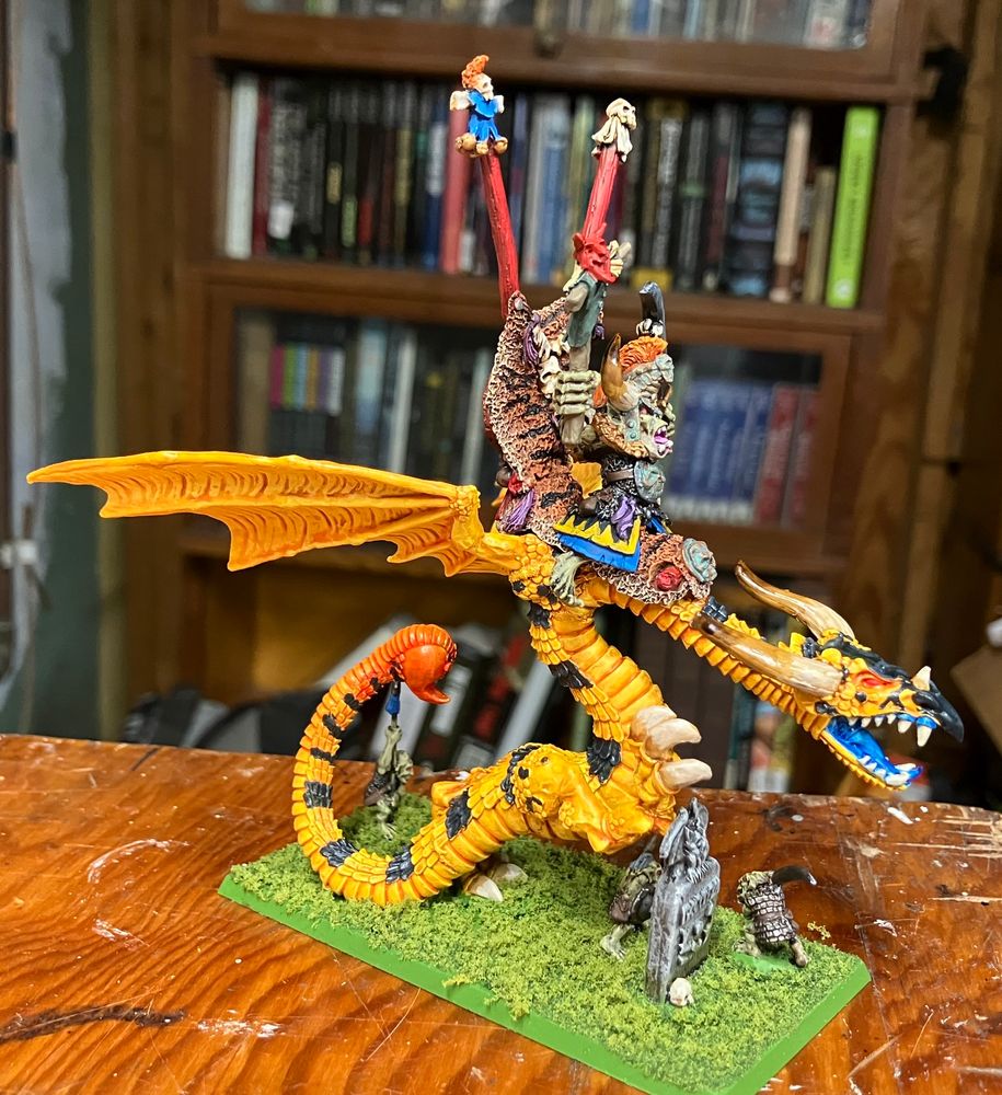 Right view of a bright yellow wyvern with black stripes and a red-tipped tail, being ridden by an orc wearing a huge horned helmet and carrying a sword and puppet. The model is on a bright green base covered with model railroad flock. 