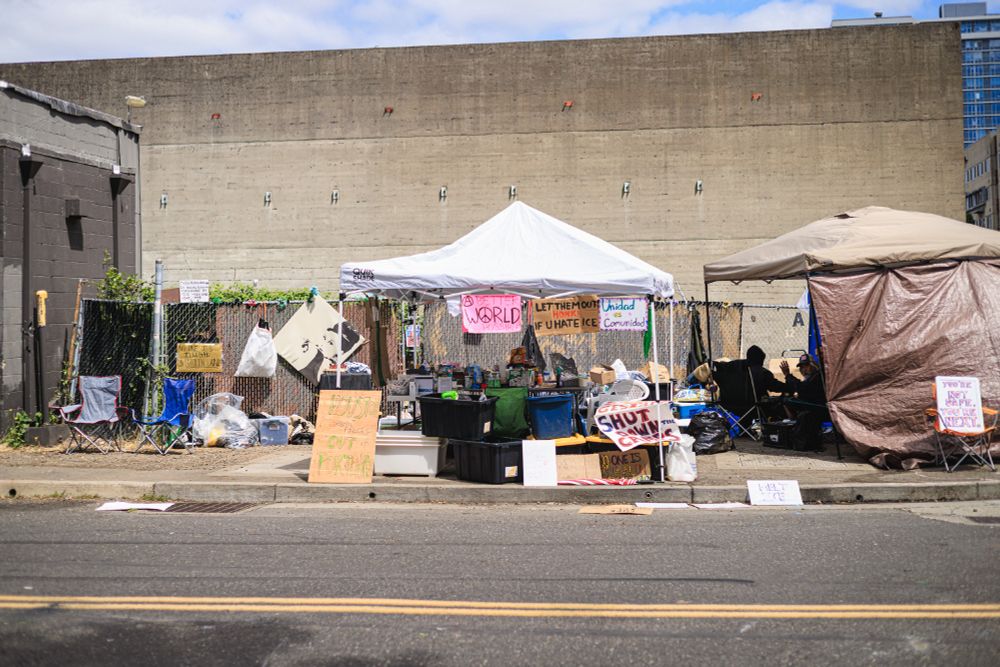 A photo of a protest encampment or occupation site set up along a street or sidewalk. The scene includes several tents (both white and brown/tan colored), camping chairs, and various supplies and belongings scattered around. There's a chain-link fence in the background with protest signs attached to it.
The visible signage includes messages like "LET THEM OUT" and references to "ICE" (Immigration and Customs Enforcement). The setting is an urban area, with concrete walls and buildings visible in the background. This type of encampment is typical of organized protests or demonstrations, particularly those focused on immigration issues given the visible references to ICE. The setup suggests this is a sustained protest action rather than a temporary gathering, given the presence of camping equipment and the organized nature of the site.
