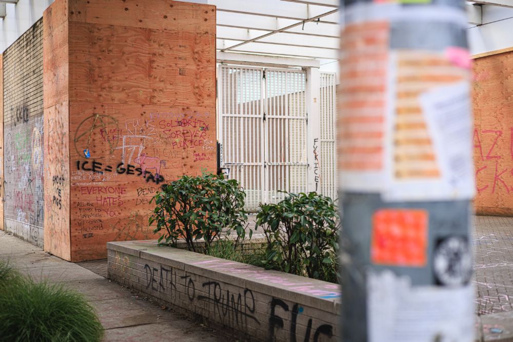 A photo of a building or structure that has been heavily covered with graffiti and protest messages. The plywood-covered walls display various writings including "ICE = GESTAPO" and other anti-ICE (Immigration and Customs Enforcement) messages. There's also graffiti that includes "FUCK NAZI FUCKS" and "SOLIDARITY FOREVER, FUCK ICE" and other political statements. The building is boarded up with plywood. There's a concrete planter with some greenery, and you can see part of what appears to be a utility pole or post covered in flyers and stickers. The overall scene suggests this is an area that has been used for political expression and protest, particularly around immigration issues, given the repeated references to ICE and related messaging.