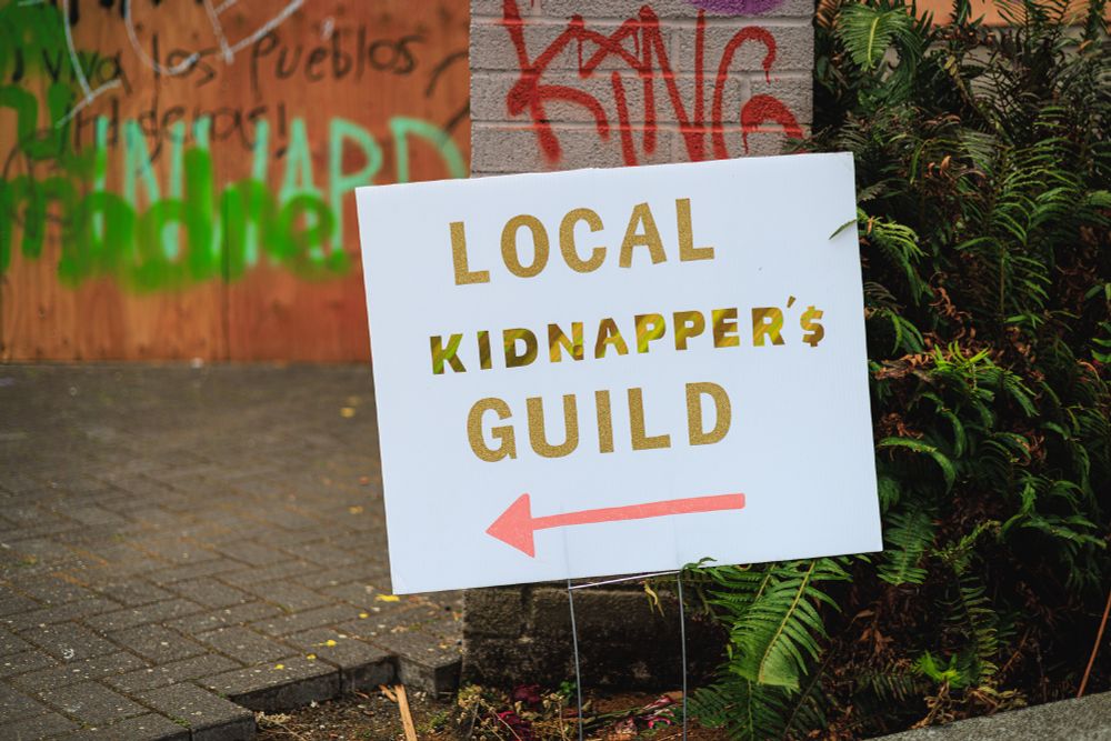 A photo of a protest sign that reads "LOCAL KIDNAPPER'S GUILD" with a red arrow pointing to the left. In the background, there's graffiti visible on walls and plywood. The sign is placed among some ferns along a sidewalk or pathway.