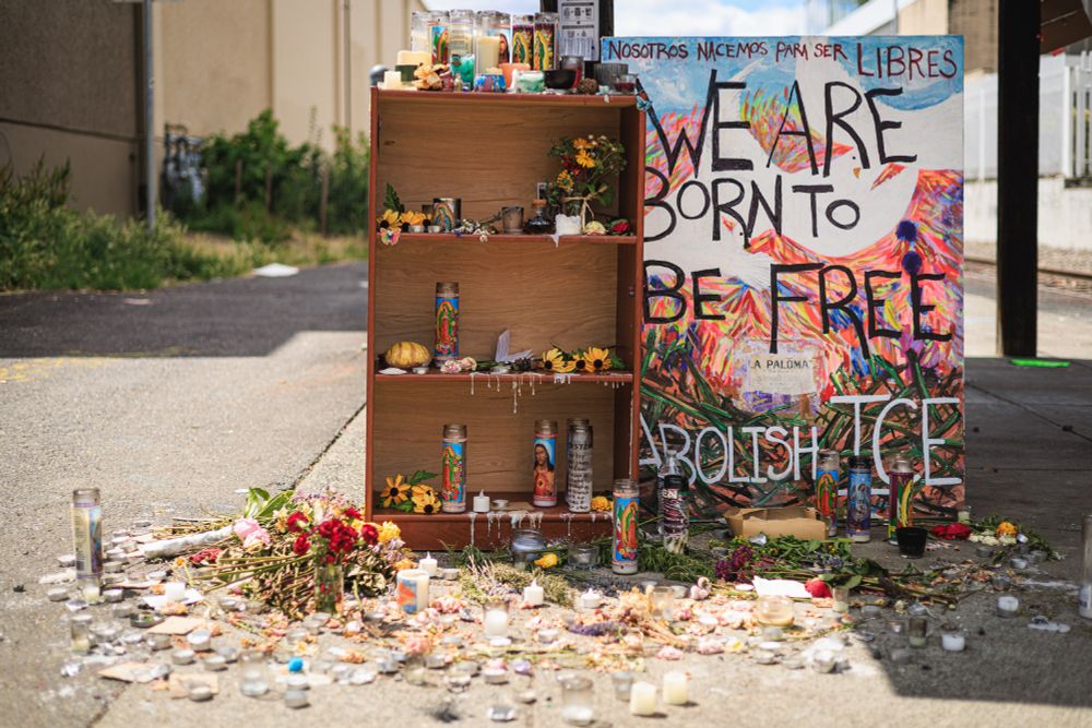 A photo of a makeshift memorial consisting of a wooden bookshelf or display case containing various items including candles, flowers, and personal objects. There's a large colorful sign or banner with text in both Spanish and English that reads "NOSOTROS NACEMOS PARA SER LIBRES" and "WE ARE BORN TO BE FREE" along with "ABOLISH ICE." The memorial is surrounded by scattered flowers, candles, and other offerings on the ground. The setting is outdoors on pavement, with buildings visible in the background. The messaging suggests it is related to immigration rights activism.