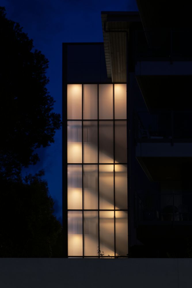 3:2, Colour, Portrait: A modern building at #night with a glass facade revealing a warmly lit staircase taking centre place. There is a mixture of glowing light and texture created by the combination of light, staircase silhouette and window frames. This shot is best understood in vertical thirds, on the left is a tree lit from behind by an evening sky. In the centre is the stairwell, on the right are grid-like window frames. This is shot over the road from the Northbridge retirement home. I’ve had several goes at this one this is the best so far. I get stared at by suspicious old folks….  #Nikon #nz #urbangaze #Auckland #aotearoa #UrbanPhotography #Glow #BlueskyArtShow