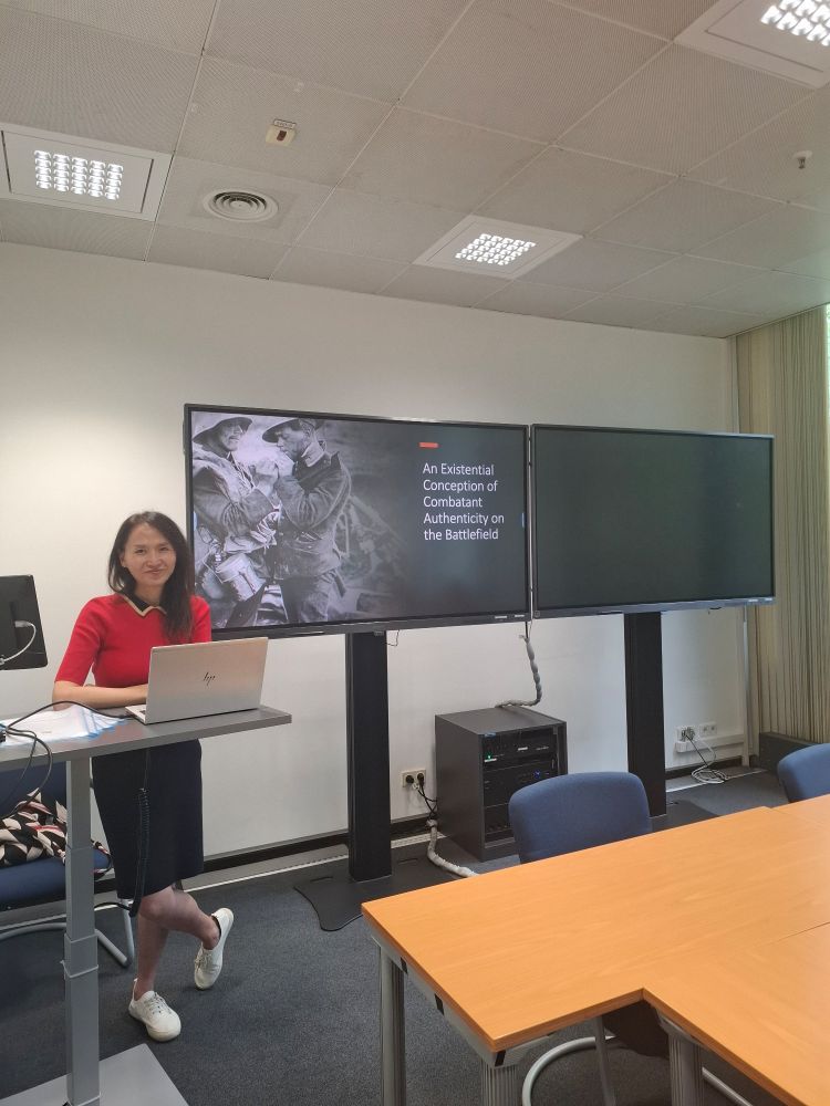 A person standing at a desk in front of a slide reading "an existential conception of combatant authenticity in the battlefield"