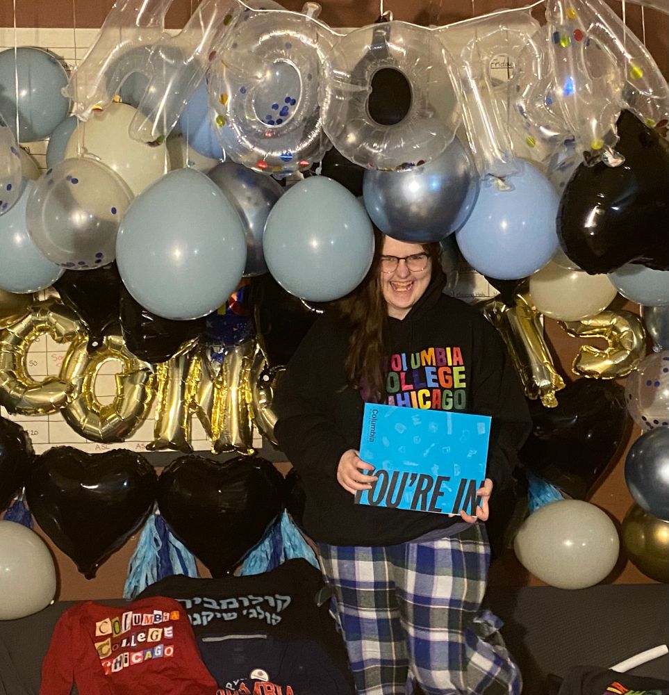 Caucasian young woman with long brown hair, glasses, and a massive smile in a Columbia College Chicago hoodie holding a blue folder that says You're In. She has balloons that are a mix between baby blue, blue grey, silver, gold, black hearts, Congrats, and Hooray surrounding her.