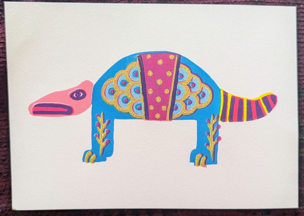 A rubber block reduction print of an alebrije armadillo. Reference is a wooden figure I purchased second hand many years ago. The body is blue, the head pink, and the tail yellow. the head has one eye and part of a down turned mouth in purple. The tail is striped between yellow, red, and purple. The body design features yellow scales filled with white and small red circles. In the middle is a red stripe with yellow polka dot design, and the legs have yellow vines moving up with red berries.