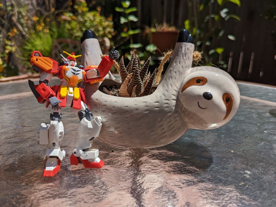 Gunpla model posed making a thumbs up in front of a sloth planter.