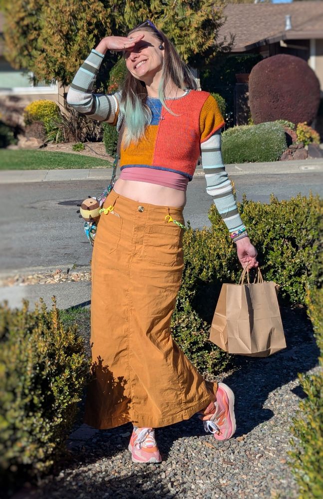 Photo of peyerate (she/they) smiling and posing for the camera with one hand blocking the sun from her face. They are wearing a colorful outfit with a self-knit colorblock tee, a striped long sleeve underneath, a mustard colored long skirt, and pink & orange sneakers.