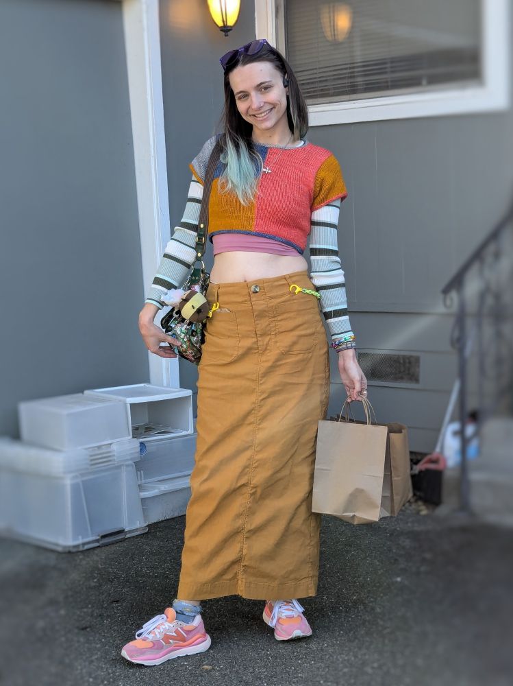 Photo of peyerate (she/they) smiling for the camera. They are wearing a colorful outfit with a self-knit colorblock tee, a striped long sleeve underneath, a mustard colored long skirt, and pink & orange sneakers.