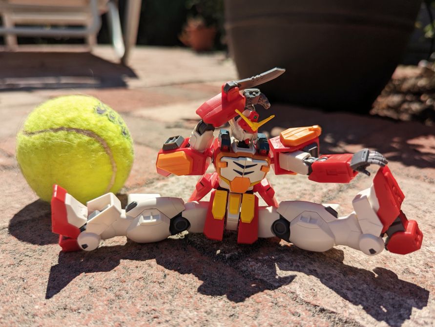 Gunpla posed in front of a tennis ball doing the splits and stretching towards one of their legs.