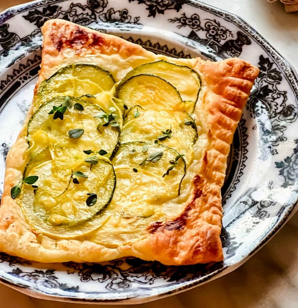 A rectangular puff pastry tart topped with thin slices of zucchini, melted cheese, and fresh herbs, served on a decorative black and white plate.