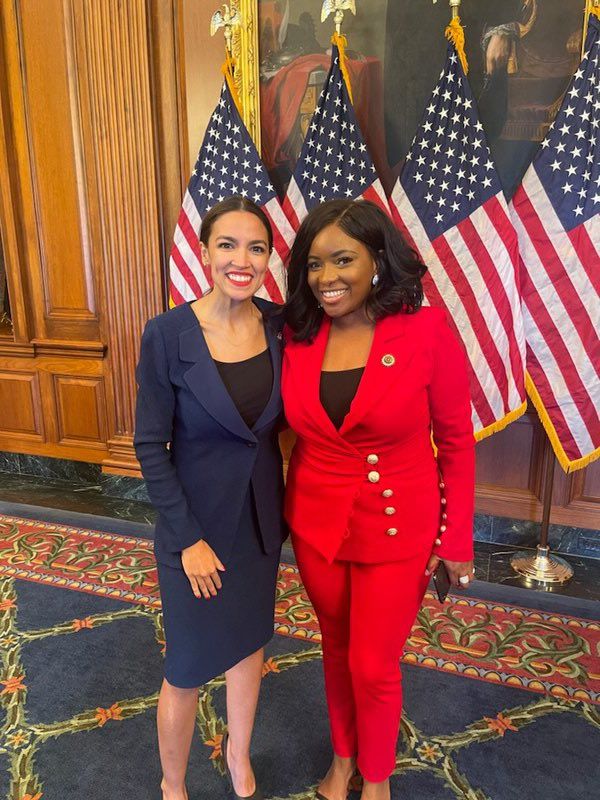 AOC and Jasmine Crockett stand smiling in front of four American flags and a wood-paneled wall with framed paintings. One wears a navy suit and the other a bright red suit with gold buttons. They are posing together on a patterned carpet. 