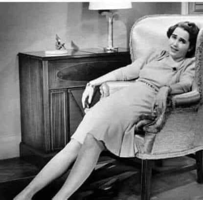 A woman in a dress lounges sideways in an armchair, appearing relaxed with her arms draped over the armrests and her legs extended out in front of her. A side table and lamp are in the background.