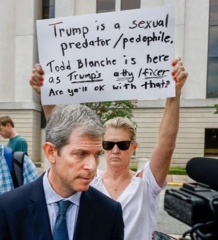 A woman wearing sunglasses holds a protest sign accusing Trump of being a sexual predator and pedophile, and mentions Todd Blanche as Trumps attorney or fixer. A man in a suit walks in the foreground outside a building.



