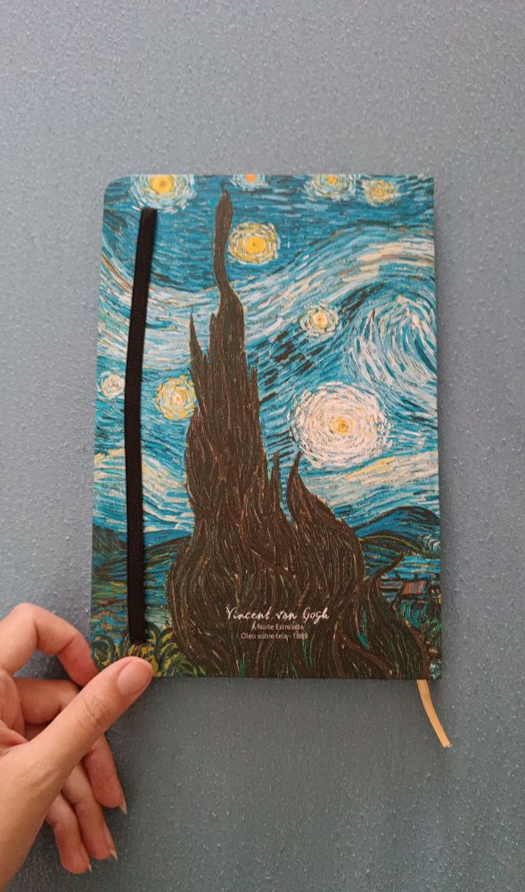 parte de trás de um caderninho. tanto a capa quanto atrás tem a reprodução da obra a noite estrelada do van gogh. tem também um elástico preto que vc pode passar na frente para manter o caderno fechado e um fitilho amarelo dentro para marcar página. minha mão está tocando na parte debaixo do caderno.