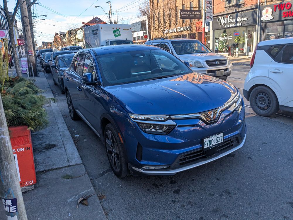 a blue vinfast VF8 parked on a street. it's a generic-looking medium-sized SUV with ontario green vehicle plates. it's got large headlights reminiscent of a dacia duster, a body-color grille that cuts across those headlights and joins in the middle below the V-shaped logo, fake air vents on the side next to the fog lights, and a real radiator vent in the middle.