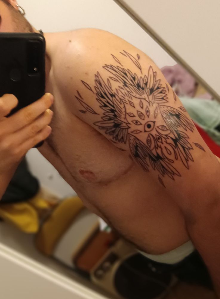 Fotografía de perfil sin camiseta en la que se ve mi brazo recién tatuado, el tatuaje es un serafín, un tipo de angel que tiene muchas alas y ojos. Está perdiendo plumas como en un estallido y los ojos están asustados