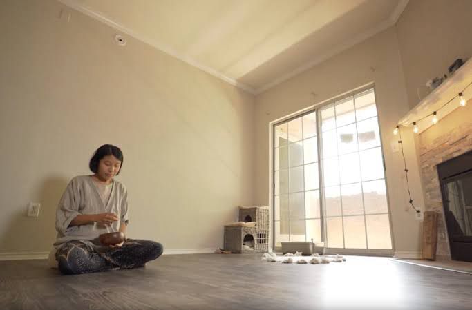 mulher sentada no chão em uma sala quase toda vazia, que contem apenas uma casinha pro gato dela