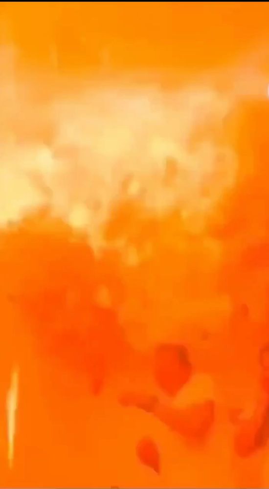 no vídeo vemos um casal hétero, num chá revelação decorado com balões pequenos nas cores roxa e amarela. ao meio, encostado no teto, um balão preto flutua e o homem tenta estourar o balão preto flutuante com uma vareta em chamas. ao fim, uma explosão acontece. no print se vê o momento da explosão onde uma labareda encobre a tela numa massa em tons diversos de laranja, podemos notar o casal ao fundo, corpos retraídos da explosão, totalmente encobertos pela cor laranja tal qual um negativo da kodak saído diretamente do inferno