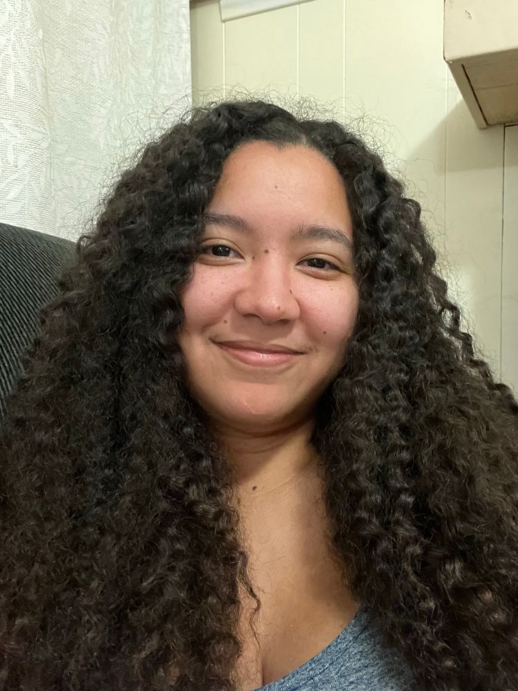 Ali (light tan skin) with brown hair that looks crimped, smiling 