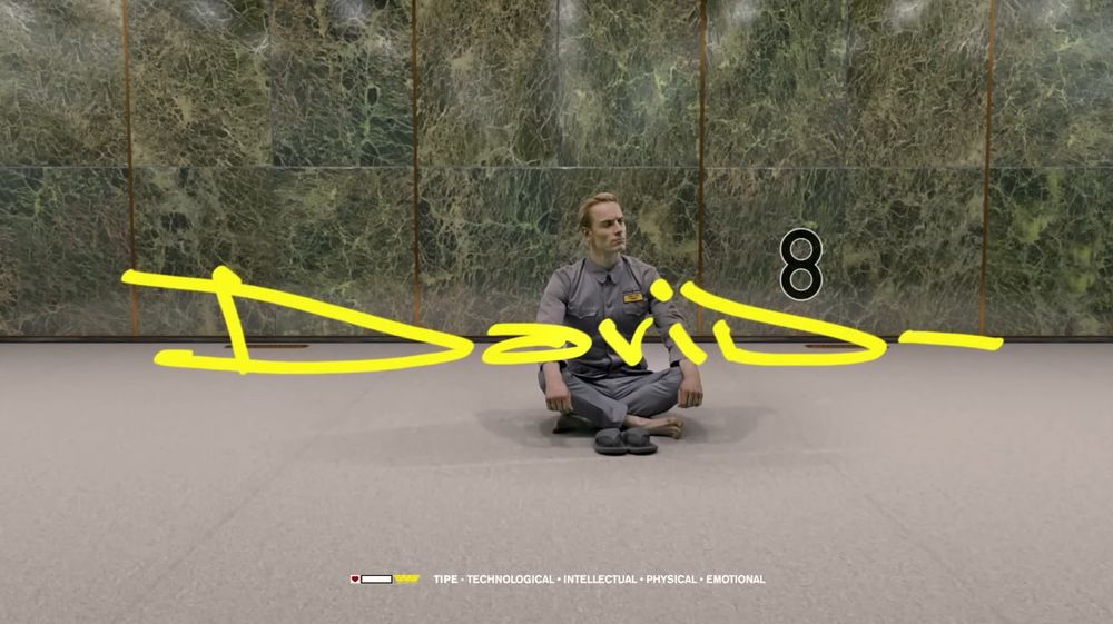 a still from Happy Birthday David, a short film directed by Johnny Hardstaff for Scott Free Productions in promotion of PROMETHEUS (2012).

David (Michael Fassbender), a synthetic human, sits cross-legged, slightly in an empty and unassuming room.

Appearing in front of him is what appears to be his own signature and indeed his logo, a basic but human and stylish wide thin script in stark yellow, with a very boring black 8 above the last d in 'David' to represent which generation of synthetic he is.