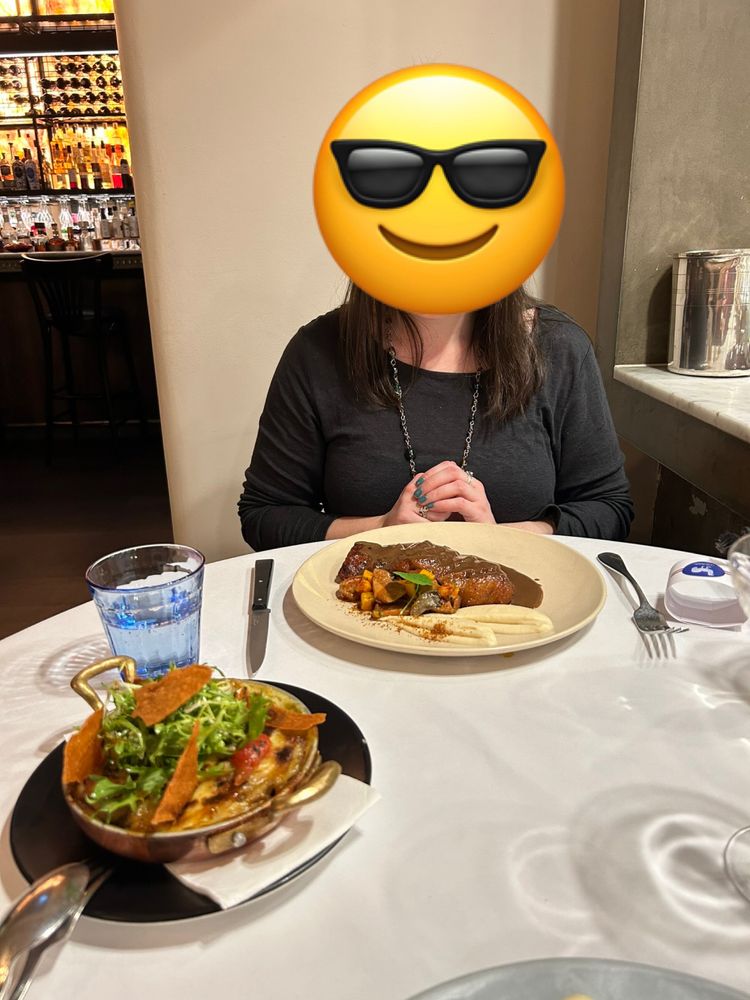 An anonymous woman sitting at a white-cloth dining table. Directly in front of her is a plate of carved duck breast covered in a luxurious dark brown sauce, alongside some cubed vegetables and a precise slick of glossy mashed potato.

In the centre of the table is a serving dish containing duck leg gratin.