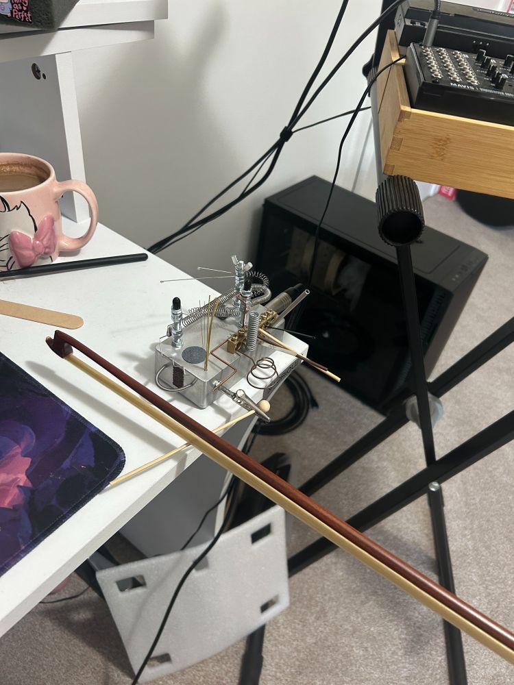 Bowing a tiny wooden mallet clamped by a metal grip attached to a metal box with lots of springs and different sized wires coming out (noise box). On edge of white desk. 
