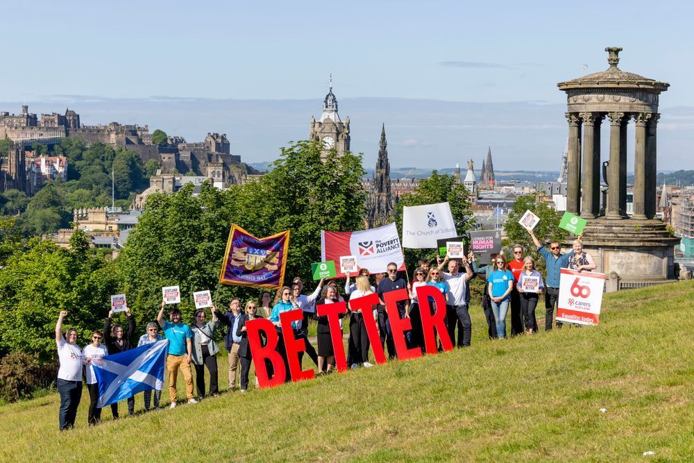 Photo of a group of people holding up protest signs and banners. At the front the word 'better' is spelled out in large red capitals, and in the background is a landscape of Edinburgh.