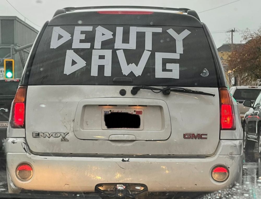 A GMC Envoy XL with DEPUTY DAWG written in white tape across the entirety of the rear window.