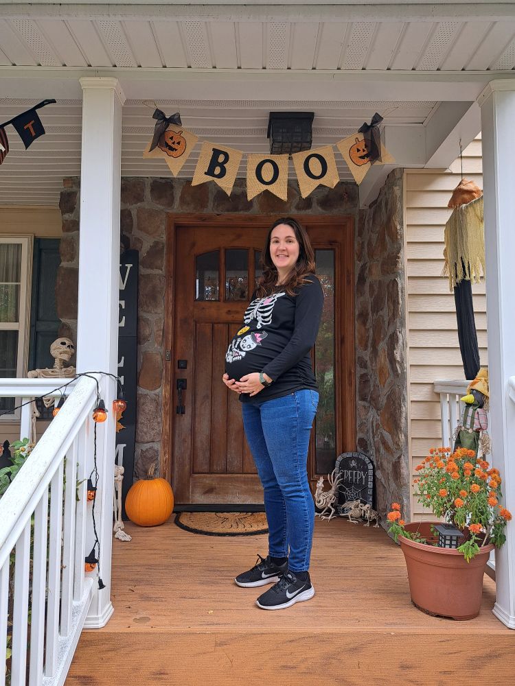 Me super pregnant in a skeleton halloween shirt holding my huge gut on my porch decorated with halloween decorations 