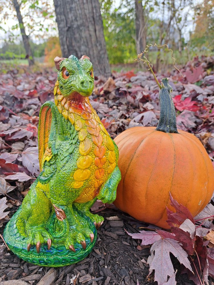 Facing forward, Finished Windstone Paint your own keeper dragon with a green body, yellow to red gradient wings and belly, orange eyes, and copper horns and claws. Next to a small orange pumpkin on brown mulch against a reddish maple leaf background.