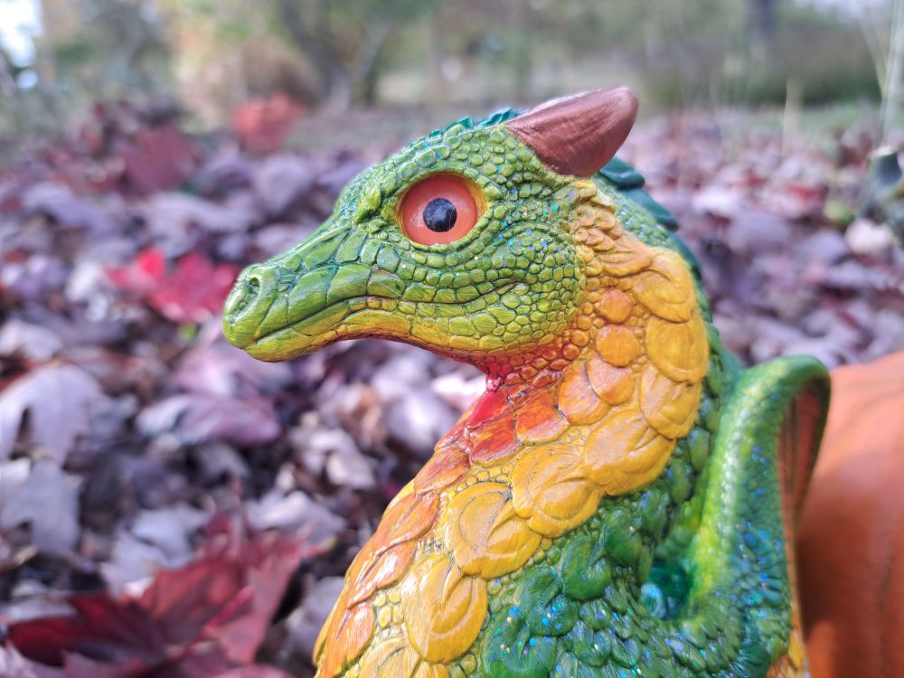 Close up of face facing left, Finished Windstone Paint your own keeper dragon with a green body, yellow to red gradient under the chin, orange eyes, and copper horns. against a reddish maple leaf background.