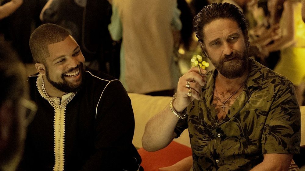 O'Shea Jackson Jr. with very short hair and a beard in a black coat smiling as Donnie Wilson and Gerard Butler with slicked back hair, a beard and a half open button down shirt holding a drink as Big Nick from Den of Thieves 2: Pantera.