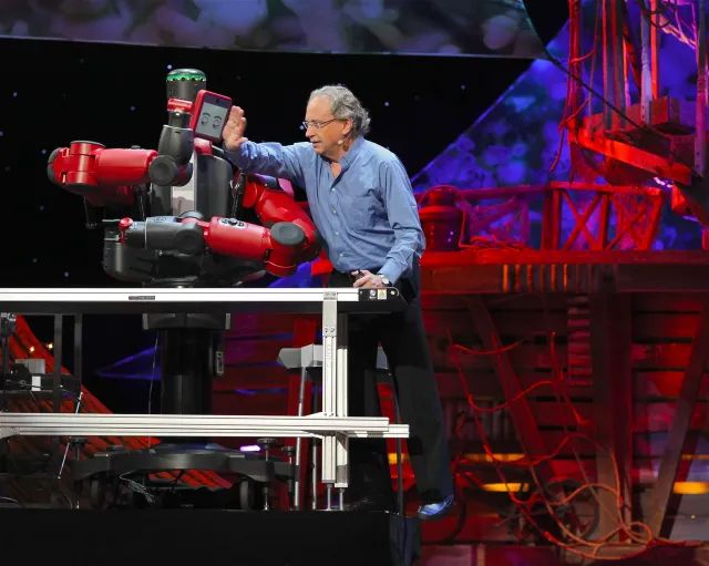 Rod Brooks on stage pushing the Baxter robot which is sitting on a table.