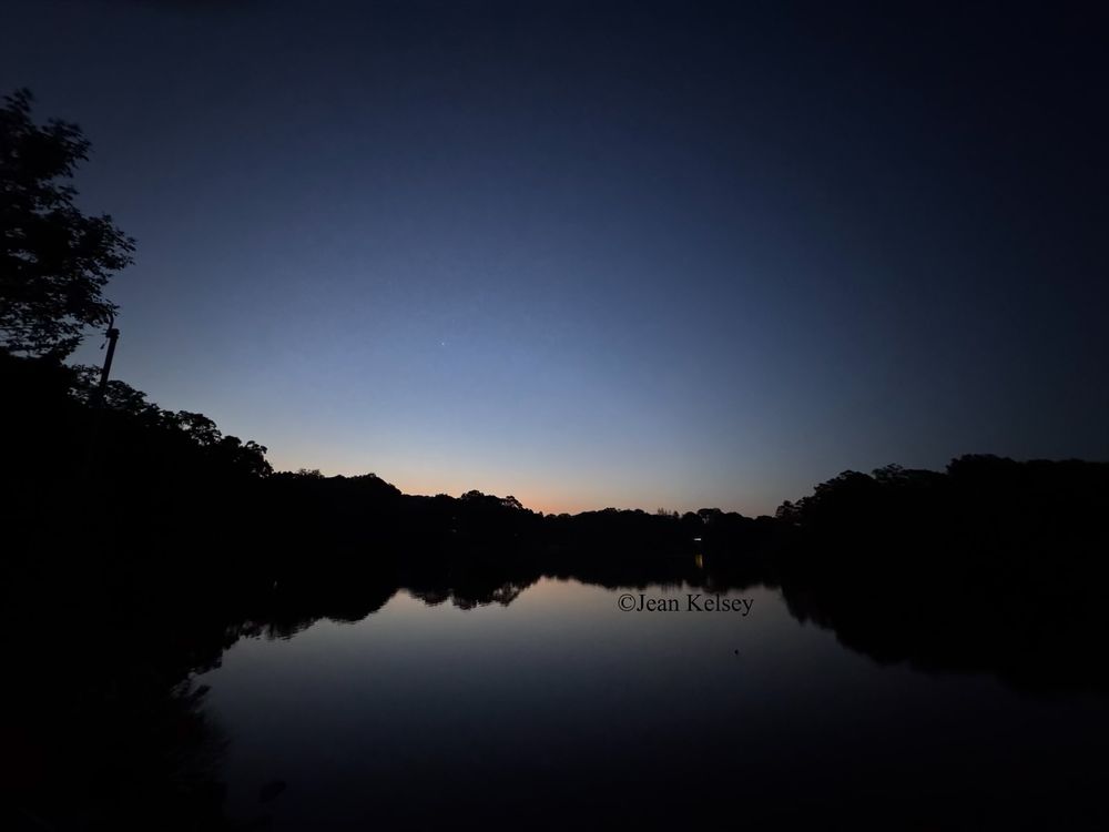 First light of the day with clear skies. Over the trees in the distance a bit of color begins to show and there is a halo of light over the trees in the distance. The water is calm reflecting the sky and landscape.
