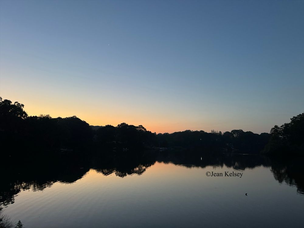 As the sun rises higher in the sky an orange glow is seen over the trees in the distance. The sky is blue and reflects on the calm waters along with the landscape.