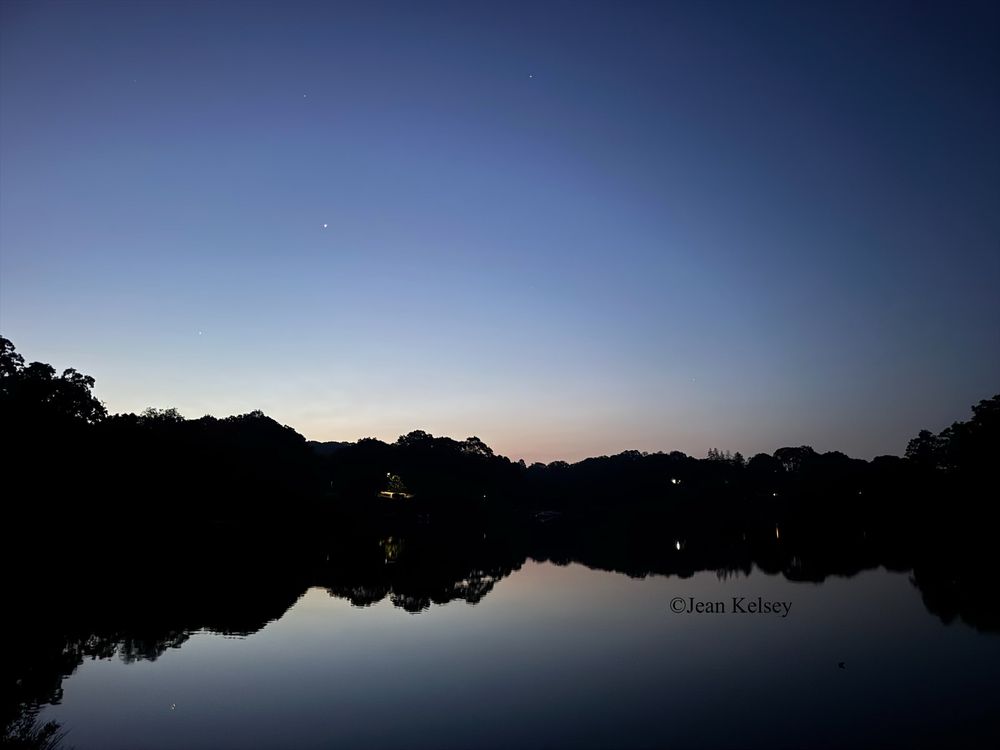 First light of the day with clear skies. Over the trees in the distance is bright light and a touch of orange color. The water is calm reflecting the landscape and trees. Lights from houses and streets dot the landscape. 