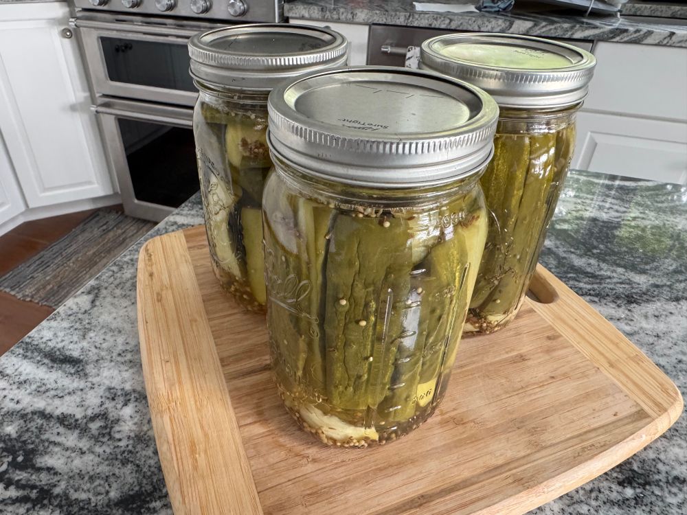 Three jars of garlic dill pickles resting on a wooden cutting board on a counter top. 