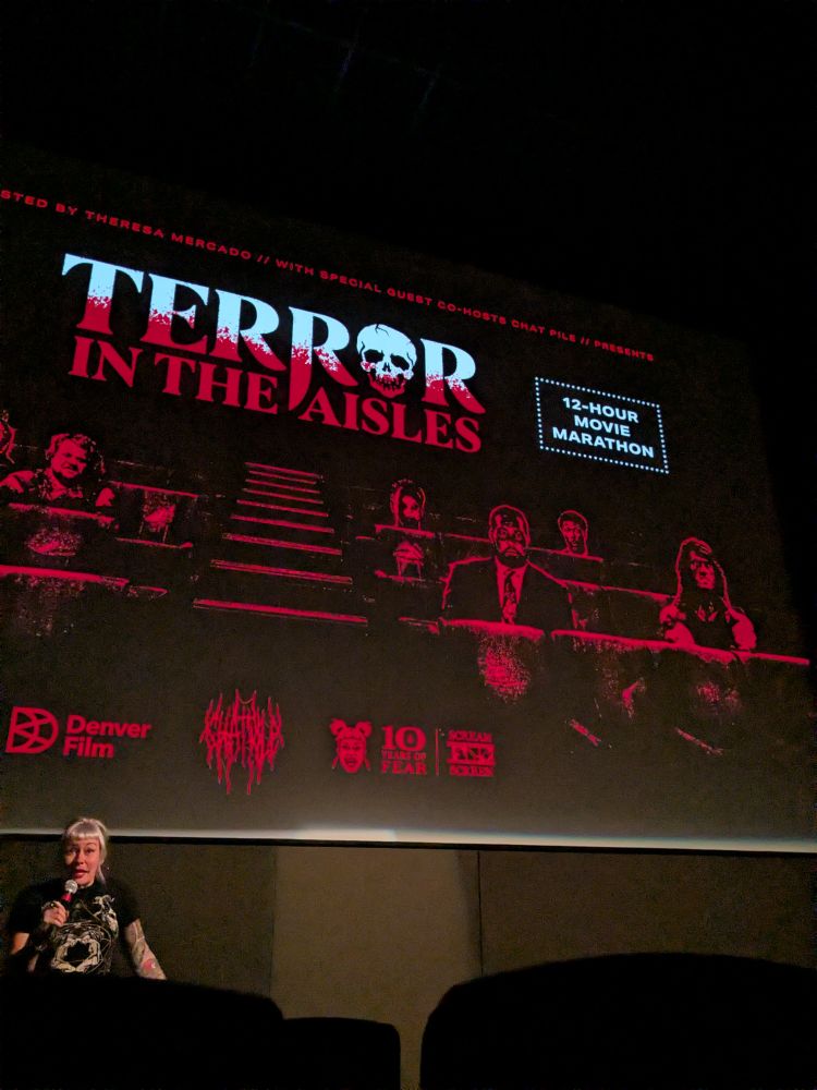 Movie theater screen advertising "TERROR IN THE AISLES: 12-HOUR MOVIE MARATHON" Teresa, who organized and is emceeing the event, is standing in front of the screen holding a microphone.