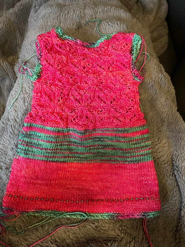 A baby tunic with scraggly ends lays flat on a grey, fuzzy blanket. The top half of the tunic is a flowery lace pattern in bright pink. Below that is a wide horizontal variegated green/pink stripe, followed by a wider band of pink. The arms, neck, and bottom hem are all edged with the variegated yarn in a picot stitch. 
Pattern: Sophie Tunic by Tabetha Hedrick
Yarn: Knit Picks Hawthorne Fingering in Parkrose, and Hazelwood
