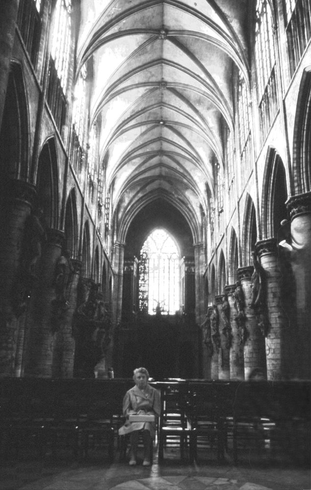 A lone woman sits quietly at the front of a cathedral’s long arcade of arches and columns. The scene is dimly lit with light from above. The seat to her left glows with the light of the cathedral’s large gothic arched west window.